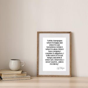 May include: A framed print with a quote about drinking champagne, in a wooden frame. The quote is in black text on a white background. A stack of books and a mug are on the table in front of the print.