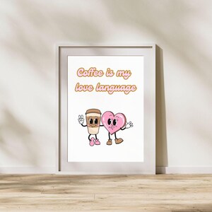 Op de afbeelding: Een wit ingelijst print met een roze en bruine cartoon koffiebeker en hart die hand in hand staan. De tekst "Coffee is my love language" is geschreven in roze en bruine letters.