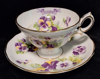 May include: A white teacup and saucer set with a gold trim. The cup features a purple and yellow floral pattern with the word "DORP" written in purple on a banner.