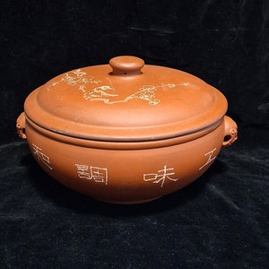 May include: A terracotta cooking pot with a lid and two handles. The pot has a white design of flowers and birds on the lid and the words "玉 蜀 味 工" on the side.