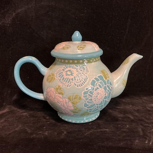 May include: A ceramic teapot with a light blue base, handle, and spout. The teapot is decorated with pink and blue floral designs and a matching lid. The lid has a small blue knob on top. The teapot is a decorative item.