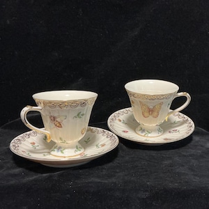 Peut inclure: Deux tasses à thé et soucoupes crème avec des bordures dorées. Les tasses et les soucoupes présentent des motifs de papillons et de fleurs. Les soucoupes ont un bord festonné. Les tasses à thé ont une anse incurvée.