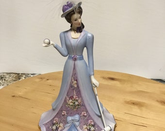 May include: A porcelain figurine of a woman in a blue and purple dress holding a white ball and a mallet. She is wearing a hat with a white feather and a floral print on her dress.