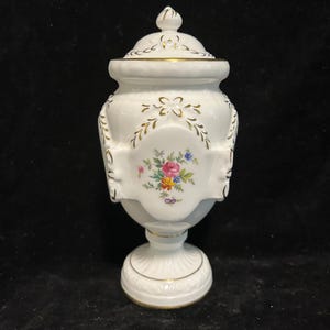 May include: White porcelain vase with a gold trim and floral design. The vase has a lid and a pedestal base.