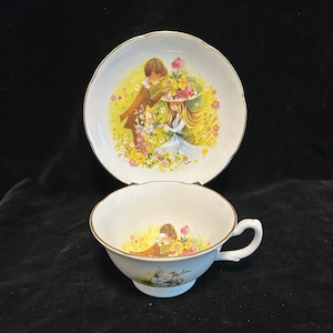 May include: A white teacup and saucer set with a floral design featuring a couple in a field of flowers. The saucer has a gold trim.