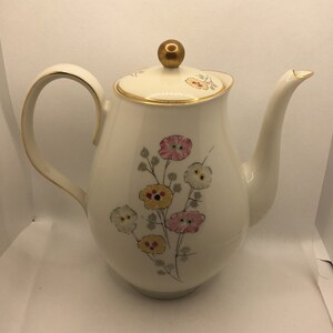May include: A white ceramic coffee pot with a gold trim and floral design. The pot has a round body, a curved handle, and a lid with a gold knob.