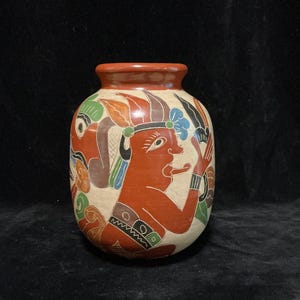 May include: A round ceramic vase with a wide neck and a smooth, reddish-brown rim. The body features a cream-coloured background decorated with hand-painted figures in red, green, blue, and black, depicting stylised human profiles and patterns.