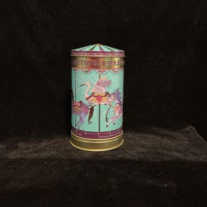 May include: A teal cylindrical tin with a carousel design featuring animals and figures. The tin has a gold base and a striped teal and purple lid. The text "FORTNUM & MASON" is printed on the lid.
