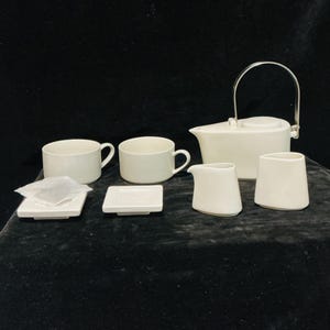 May include: A white ceramic tea set consisting of a teapot with a metal swing handle, two tea cups, two small square saucers, a creamer, and a sugar bowl. The pieces feature a smooth, matte finish and a clean, geometric design aesthetic.
