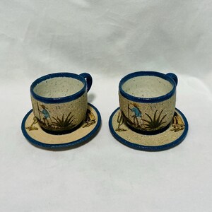 Peut inclure: Deux tasses et soucoupes en céramique avec une finition beige mouchetée et des bords bleus. Chaque pièce présente une scène peinte à la main d'un personnage en chemise bleue et chapeau de paille près d'un agave vert et d'un petit cactus.
