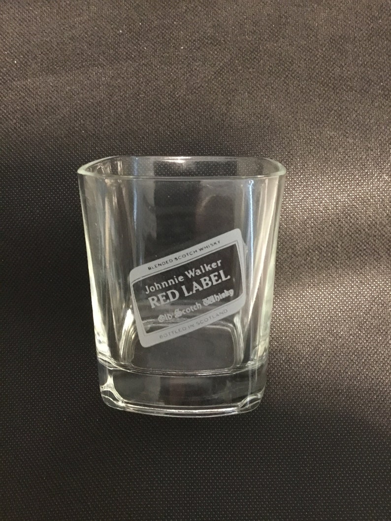 Op de afbeelding: Een helder glazen tumbler met een gegraveerd Johnnie Walker Red Label Old Scotch Whisky logo. Het logo bevindt zich in een witte rechthoek met zwarte tekst.