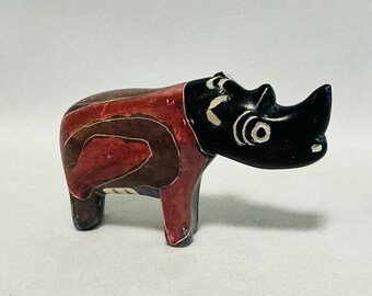 May include: A small, decorative rhino figurine. The rhino has a black head and horn, with white eye details. The body is a mix of red, brown, and tan colors. The figurine is standing on four legs and appears to be made of stone or a similar material.