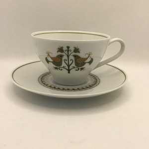 May include: A white teacup and saucer set with a green and brown bird design. The birds are facing each other and are perched on a branch with leaves.