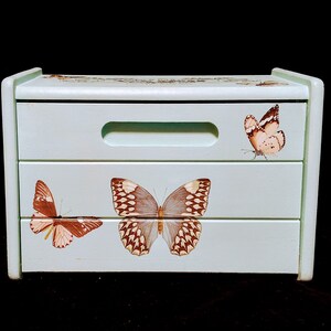 May include: A light blue wooden box with three drawers. Each drawer features a different butterfly design. The butterflies are brown and white with black markings.