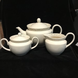 May include: A white porcelain tea set comprising a teapot, sugar bowl, and creamer. Each item features a delicate, openwork pattern around the rim. The set is presented on a dark surface.