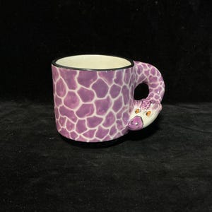 May include: A ceramic mug featuring a purple and white giraffe-patterned exterior. The handle is sculpted into the shape of a giraffe head with orange eyes and a white muzzle, extending from the side of the mug. The interior of the mug is white.