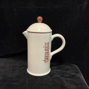 May include: Cream-coloured ceramic French press with a lid and handle. The words "La Chocolatiere" are printed vertically in brown on the side. The lid is topped with a small, round, brown knob. The spout is on the left side.