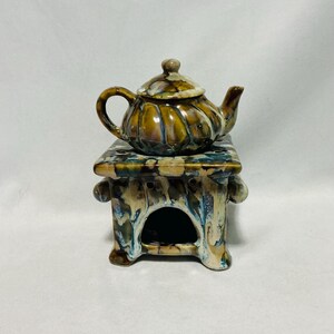 May include: A ceramic teapot with a lid rests on a decorative ceramic stand. The teapot and stand are in shades of brown, yellow, and blue. The stand has an arched opening and decorative side handles.