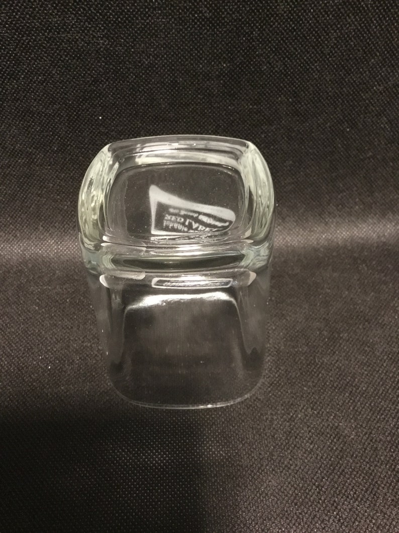 Op de afbeelding: Een helder glazen tumbler met een vierkante vorm en afgeronde randen. De bodem van het glas heeft de tekst "RED LABEL" erin gegraveerd.