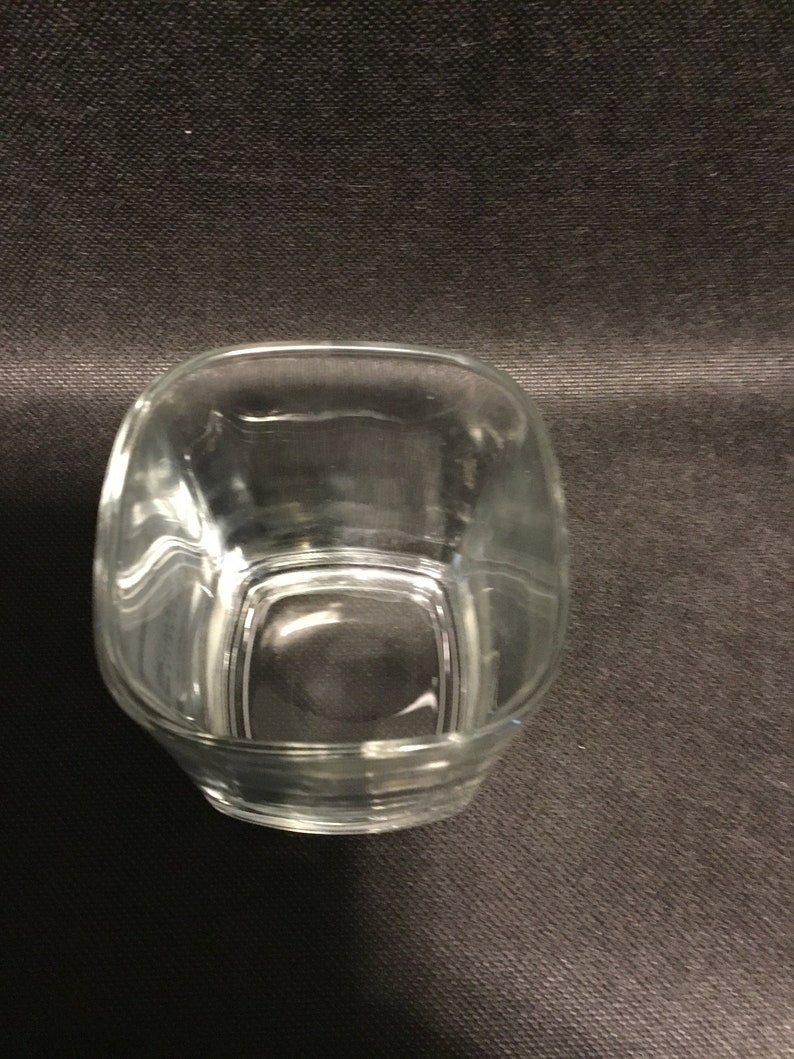 Op de afbeelding: Een transparante glazen tumbler met een vierkante vorm. Het glas is leeg en heeft een lichte kromming aan de zijkanten.