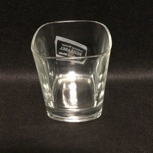 Op de afbeelding: Een helder glazen tumbler met een vierkante vorm en afgeronde randen. Het glas heeft een label met de tekst "Johnnie Walker Red Label Blended Scotch Whisky".