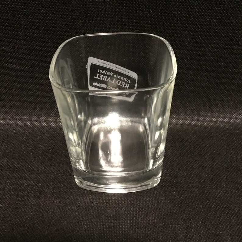Op de afbeelding: Een helder glazen tumbler met een vierkante vorm en afgeronde randen. Het glas heeft een label met de tekst "Johnnie Walker Red Label Blended Scotch Whisky".