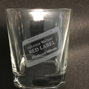 Op de afbeelding: Een helder glazen tumbler met een Johnnie Walker Red Label Scotch Whisky logo gegraveerd aan de zijkant. Het logo is zwart-wit en bevat de merknaam "Red Label" en "Old Scotch Whisky".