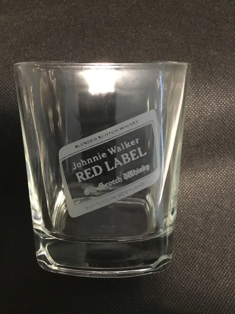 Op de afbeelding: Een helder glazen tumbler met een Johnnie Walker Red Label Scotch Whisky logo gegraveerd aan de zijkant. Het logo is zwart-wit en bevat de merknaam "Red Label" en "Old Scotch Whisky".