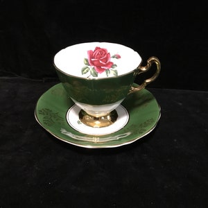 Peut inclure: Une tasse à thé et une soucoupe en porcelaine, de couleur verte et blanche, avec une bordure dorée. La tasse à thé est décorée d'une rose rouge unique avec des feuilles vertes sur fond blanc. La soucoupe a un fond vert avec des accents dorés.
