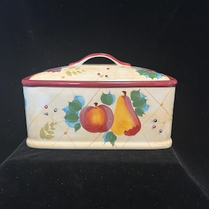 May include: A ceramic bread bin with a white background and a red, yellow, and green fruit design. The bin has a handle on the top and a red trim around the edge.