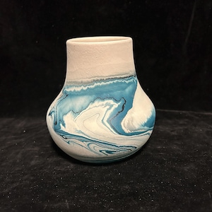 May include: A small, white ceramic vase with a blue and black marbled design. The vase has a rounded bottom and a slightly flared opening.