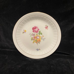 May include: White ceramic plate with a gold rim and a floral design featuring a pink rose and other colorful flowers.