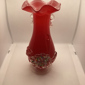 Peut inclure: Un vase en verre rouge avec un dessus à volants et des poignées en verre clair. Le vase a une base en verre clair avec un motif floral et des accents en verre coloré.