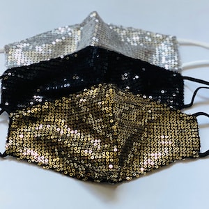 Puede incluir: Tres mascarillas faciales hechas de tela de lentejuelas. Las mascarillas están superpuestas, con la mascarilla dorada en la parte inferior, la mascarilla negra en el medio y la mascarilla plateada en la parte superior. Cada mascarilla tiene lazos elásticos para las orejas.