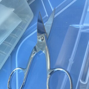May include: Shiny, silver-colored scissors with curved handles and sharp blades. The scissors are designed for precision cutting and are likely used for crafts or sewing. The blades are open, showing their sharp edges. The scissors are on a blue surface.