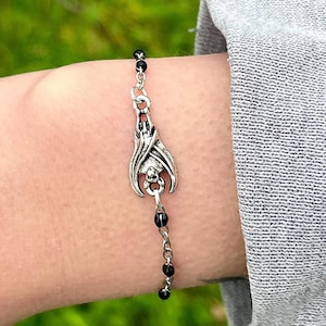 Op de afbeelding: Een zilveren armband met een vleermuisbedel en zwarte kralen. De armband zit om een pols.