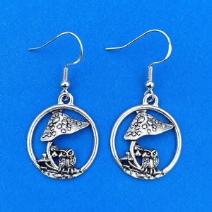 May include: Silver earrings with a mushroom and owl design. The mushroom has a white and gray speckled cap and the owls are facing each other.