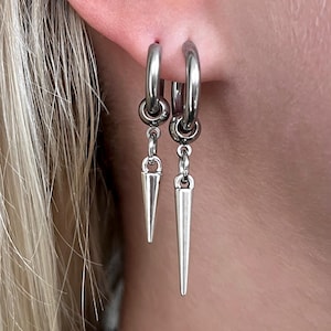 Peut inclure: Gros plan sur une oreille ornée de plusieurs boucles d'oreilles argentées. Deux créoles pendent avec des breloques en forme de cône. Une boucle d'oreille à chaîne avec un petit charme rond est également visible.