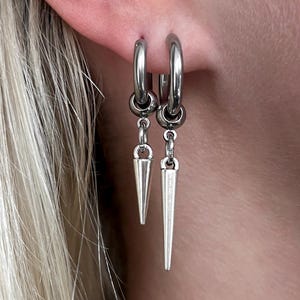 Puede incluir: Pendientes de aro plateados con colgantes puntiagudos. Los pendientes est&aacute;n sujetos a una oreja. Tambi&eacute;n se ve una peque&ntilde;a cadena plateada con un dije en forma de coraz&oacute;n.