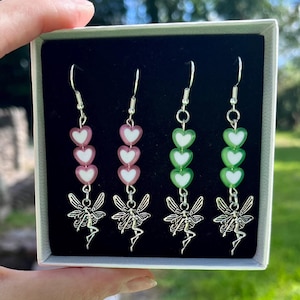 Puede incluir: Tres pares de pendientes colgantes con abalorios de hadas plateados y tres cuentas en forma de corazón. Las cuentas son rosas y verdes.