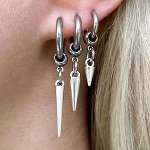 Può includere: Orecchini a cerchio argentati con ciondoli a forma di cono pendenti. Gli orecchini sono indossati su un orecchio, con più piercing visibili. I gioielli hanno una finitura metallica lucida, che riflette la luce. Lo stile generale è audace e moderno.