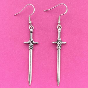 May include: A pair of silver sword earrings with a detailed design. The earrings are hanging from silver hooks.