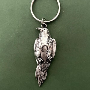 May include: Silver key chain featuring a detailed raven figurine holding a crescent moon in its talons.