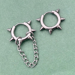 以下が含まれることがあります： パンク・ロック風のシルバーカラーのフープピアス。各イヤリングは、フープの周りに複数の円錐形のスパイクと小さなチェーンを備えています。イヤリングは、明るい緑色の背景に表示されます。