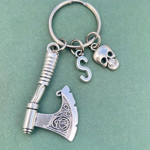 以下が含まれることがあります： 銀製のキーホルダー。ケルトの結び目のデザインが施された小さな斧、骸骨のチャーム、文字 'S' のチャームが付いています。キーホルダーは銀製のキーリングに取り付けられています。