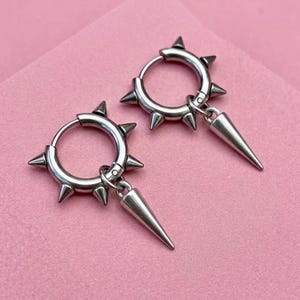Op de afbeelding: Een paar zilverkleurige oorringen met meerdere kleine stekels langs de buitenrand en een enkele, grotere, afneembare kegelvormige bedel die aan elke ring hangt. De oorbellen hebben een gepolijste metalen afwerking en een scharniersluiting.