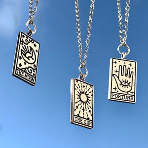 Three tarot card necklaces are pictured. From left to right they are the moon, the sun and fortune. A delicate silver chain is attached to the tarot card pendants. The pendants are rectangle shaped.