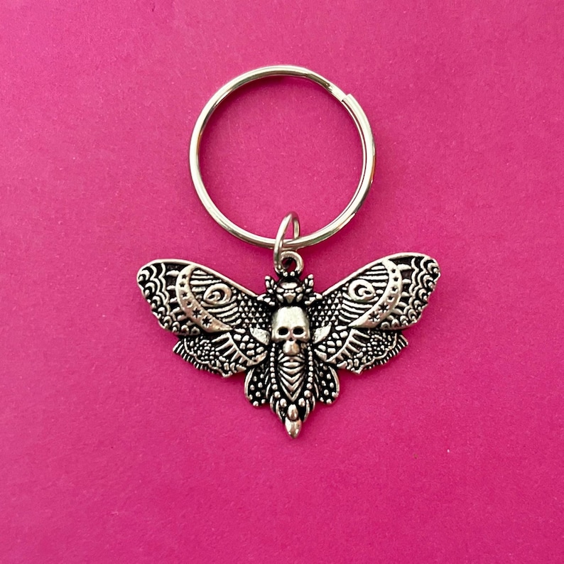 May include: A silver key chain with a detailed moth charm featuring a skull in the center. The moth has intricate patterns and a silver key ring.
