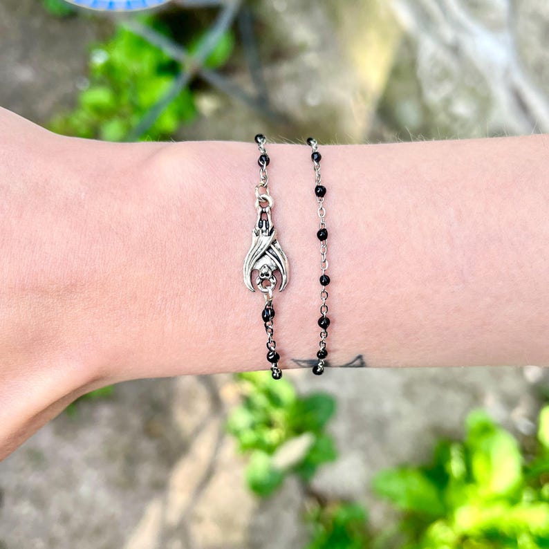 Peut inclure: Un bracelet argent&eacute; orn&eacute; d'un pendentif chauve-souris et de perles noires. Le pendentif chauve-souris pr&eacute;sente des d&eacute;tails complexes. Le bracelet est port&eacute; sur un poignet. L'arri&egrave;re-plan est flou avec des feuilles vertes.