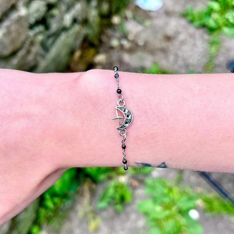 May include: A silver bracelet featuring a crescent moon charm with embedded crystals. The bracelet has a delicate chain with small black beads. The charm is the focal point, adding a celestial touch to the jewelry.